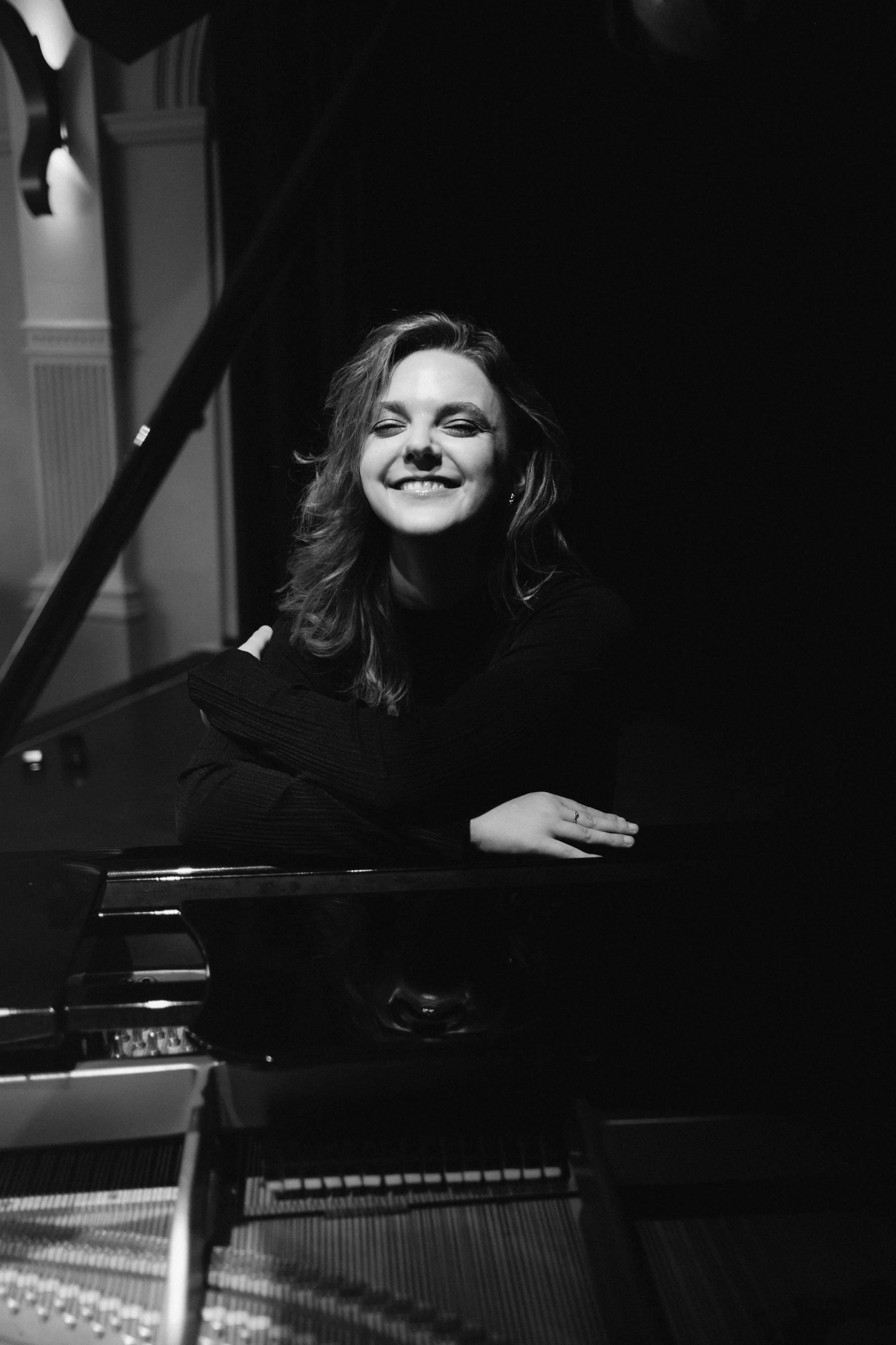 Black and white photo of a woman with wavy hair smiling and leaning on a grand piano inside a room with decor.