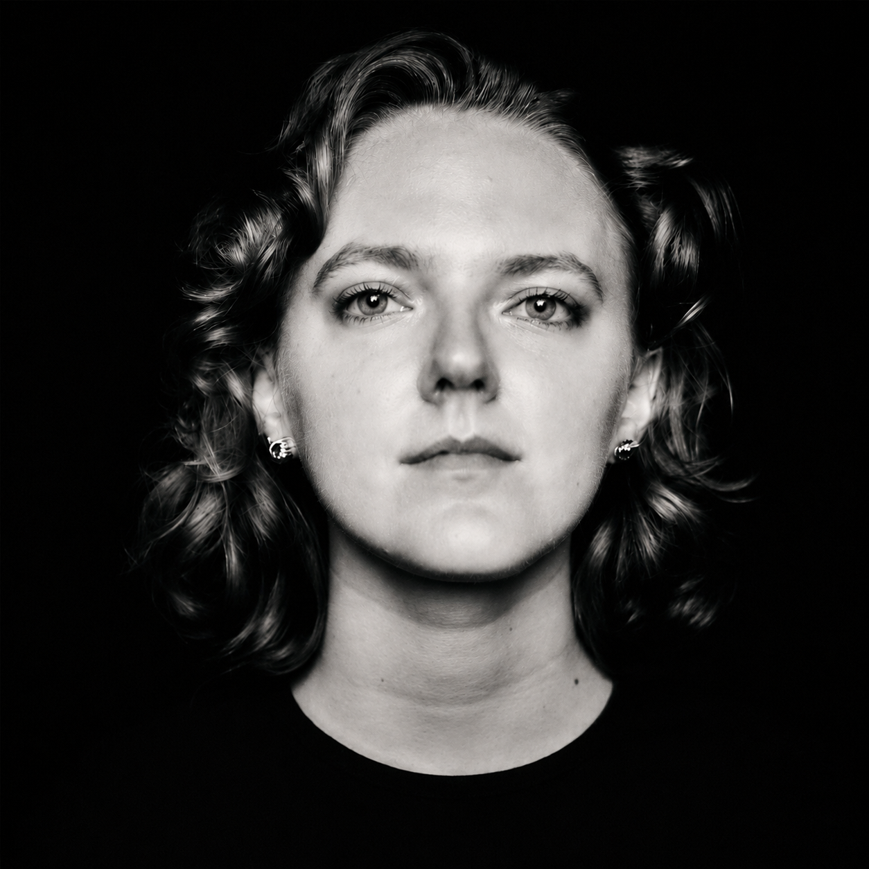 Black and white portrait of a woman with short, wavy hair and earrings, looking directly at the camera against a dark background.