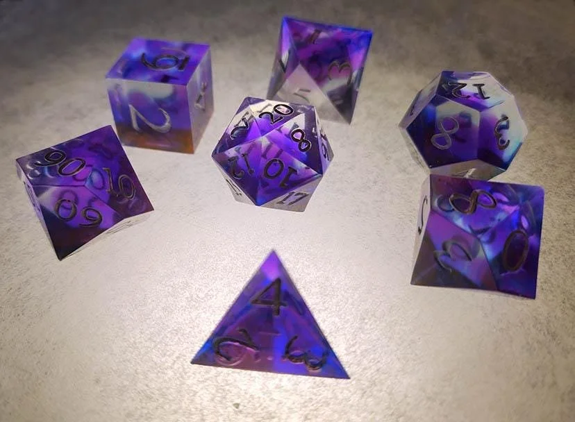 Detailed photo of JoaraCrafts' Purple Nebula dnd dice set Focused on the D20 with the bright purple and blue swirls inside the clear resin, the white numbers seemingly looking black when placed over the light surface. All dice are noticeably handcraf