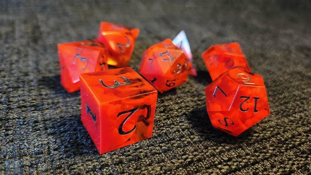 Detailed photo of JoaraCrafts' Clear Ember dnd dice set focused on the D6 and D12 where you can clearly see the red swirls inside the orange dice, while the numbers are visibly black over a black surface. All dice are noticeably handcrafted.