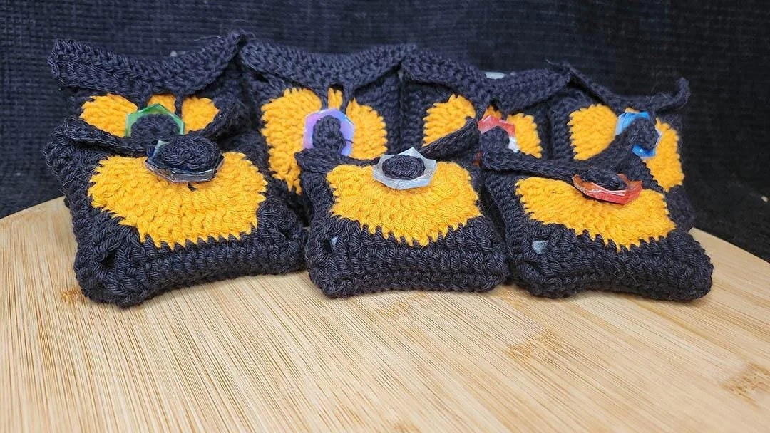 A photo of JoaraCrafts Anti-Waste Mystery Dice Pack. In this photo you can see several full handcrafted crochet black pouch with the orange hexagon representing the JoaraCrafts logo finished and a Anti-Waste resin button. The bag is set on a wooden p