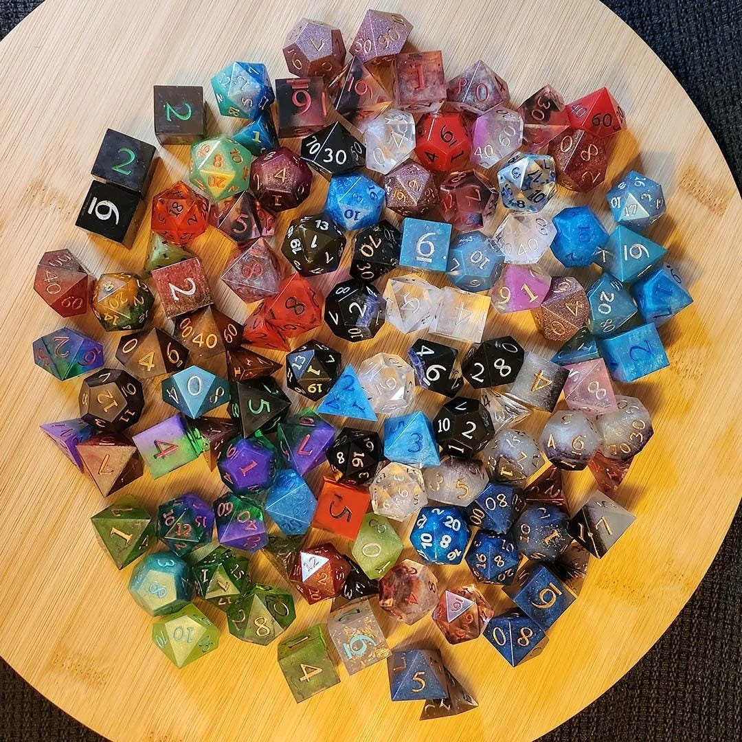 In this photo there's a representation of multiple variants of possible randomly selected mystery dice available in this packs. There's red, blue, pink, black, clear dice. All dice are displayed on top of a wooden background so it's possible to see a