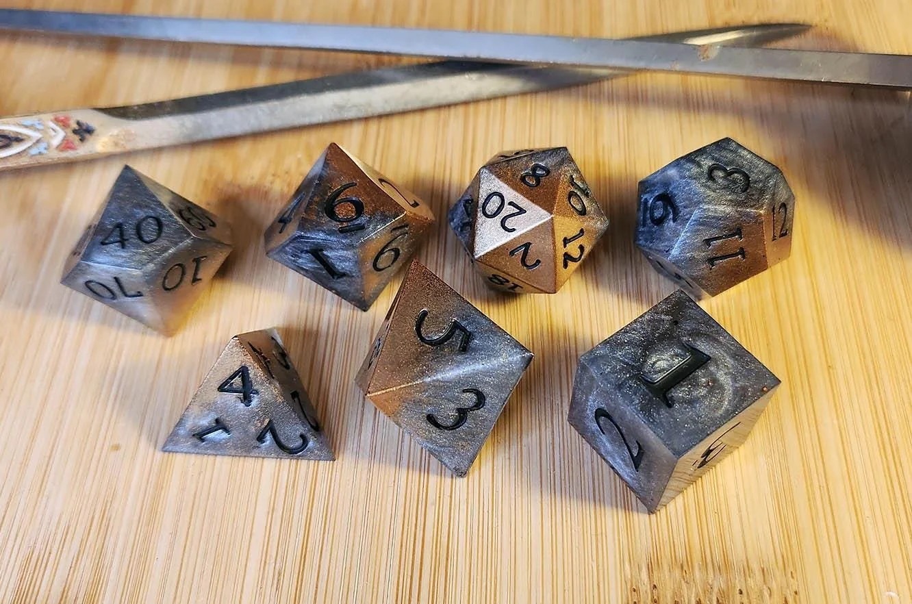 A selection of multi-sided dice in metallic shades of silver, bronze, and gold. The dice are displayed on a wooden surface, with the numbers clearly visible. Suitable for role-playing games and other tabletop activities.