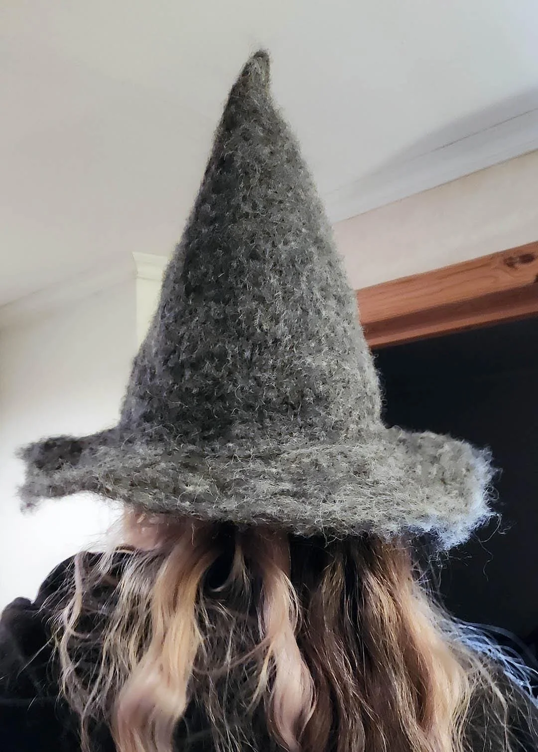 A grey, fuzzy, pointed witch or wizard hat with a wide brim. This handcrafted hat is handmade using a technique named needle felting where the crafter delicately stabs wool into a sponge and shapes the piece over time.