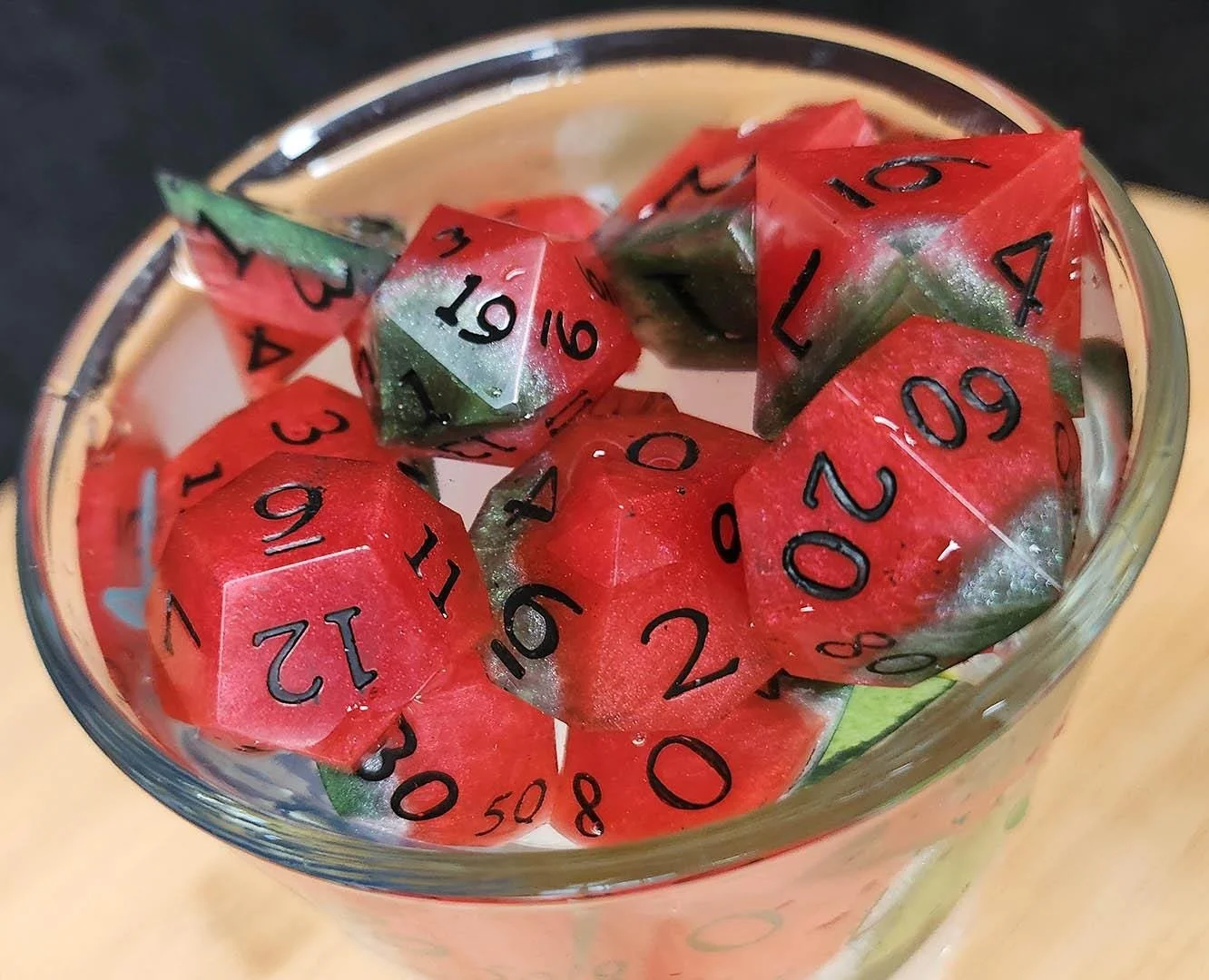 A glass filled with water containing multiple bright red, green and white watermelon styled dice, submerged in water.