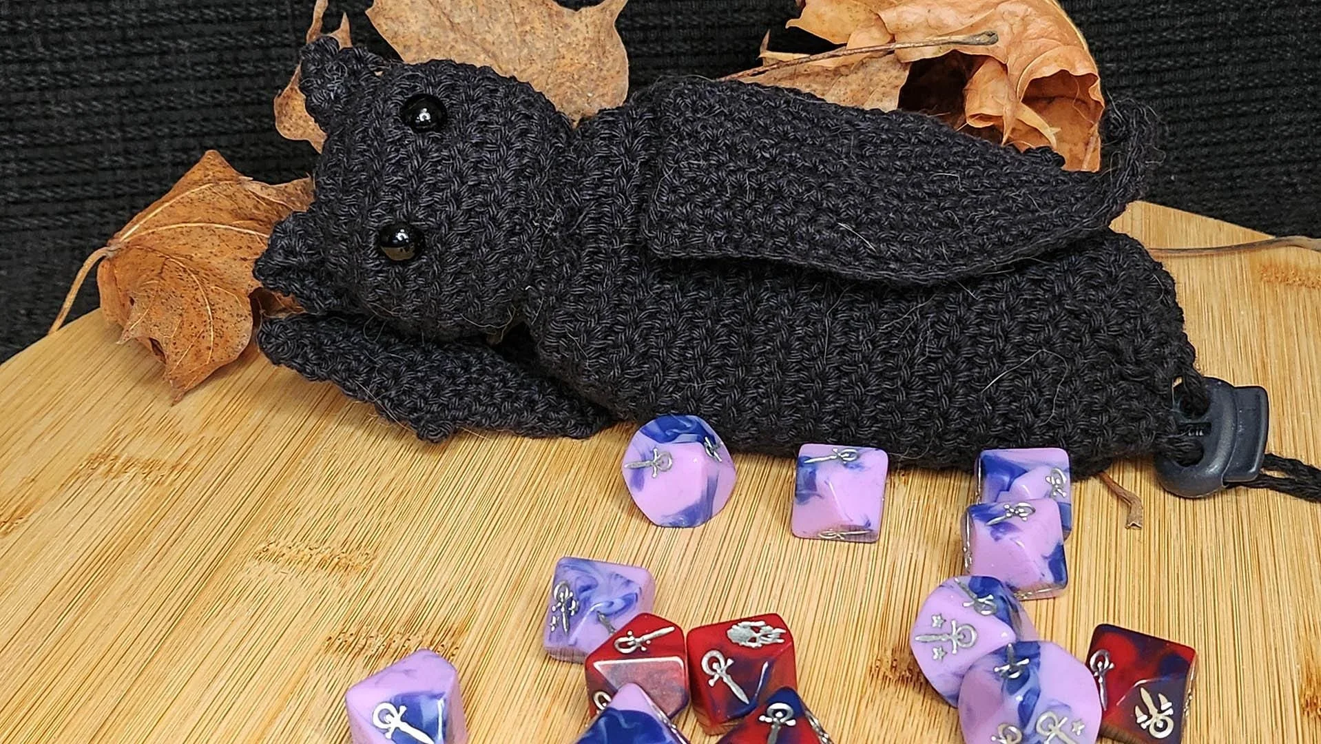 A black crocheted bat-shaped dice bag with black eyes and wings, lying on a wooden surface. Several purple and blue dice with silver symbols are scattered around the bag. The dice bag is approximately 20 cm long.