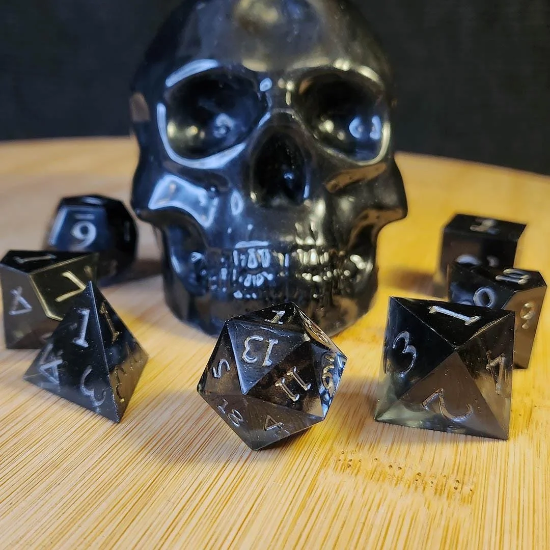May include: A shiny black skull ornament with a collection of black and silver dice. The dice are in various shapes, including a pyramid, a cube, and a 20-sided die. The numbers on the dice are silver.