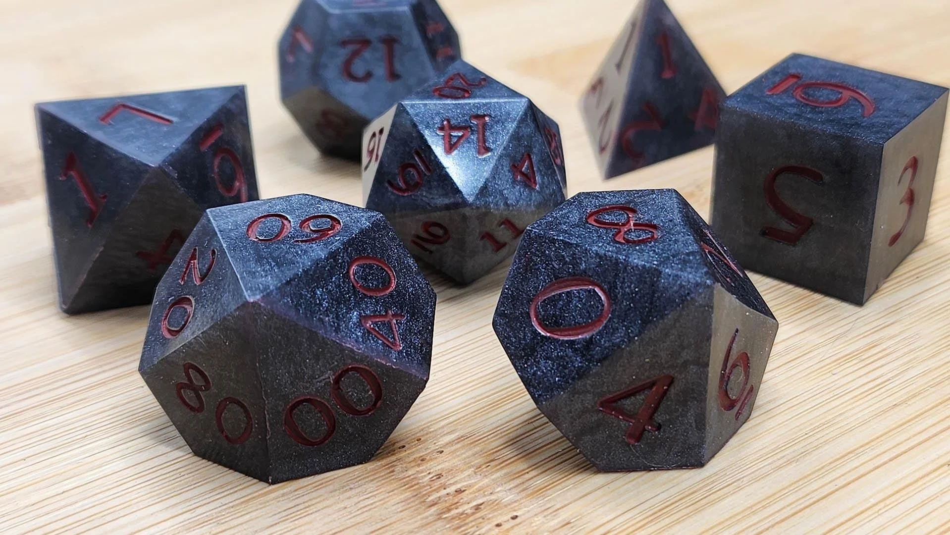 A set of seven polyhedral dice with a dark metallic marbled black and gray surface and red numbering, arranged on a light wooden surface.