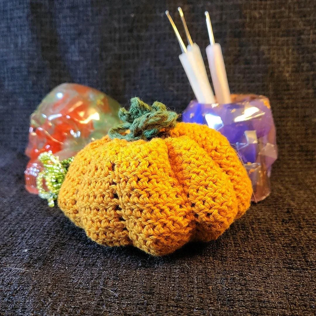 A crocheted orange pumpkin with a green stem. The pumpkin is sitting on a black surface with a purple anti-waste geometric skull and a red, green and white anti-waste skull decoration in the background.