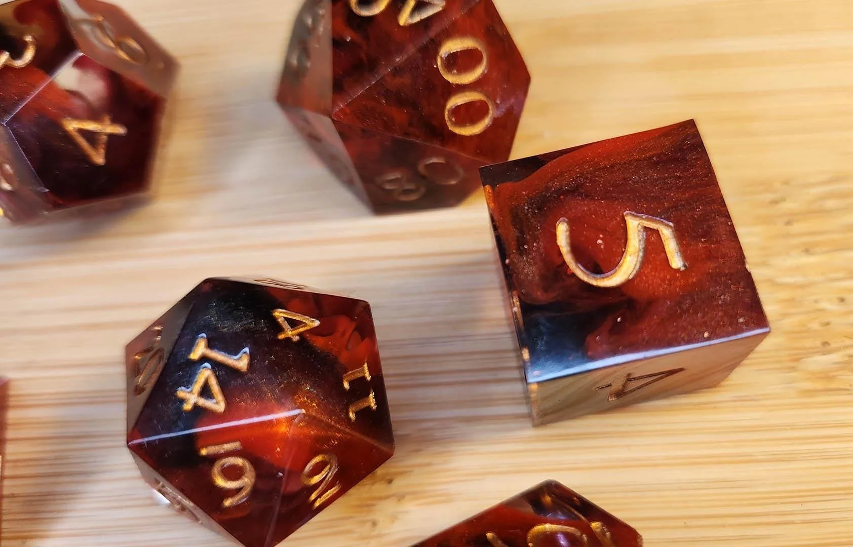 The photo shows JoaraCrafts Crimson Shadows dnd dice set. A dice set with black and shimmering orange crimson red swirls seen through a body of clear and ruby inked resin. The numbers are colored in a bright golden color and the dice are set on a woo