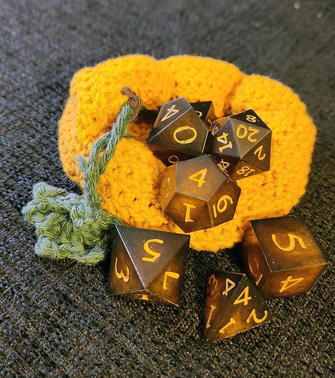 Photo of JoaraCrafts' handmade orange and black DnD dice set being removed from a crochet pumpkin dice bag. Both the polyhedral dice and the crochet bag are artisan handcrafted pieces that complement each other perfectly. The vibrant autumn orange to