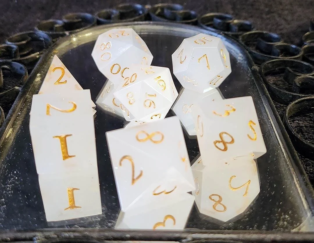 A collection of white polyhedral gaming dice with gold numbers, including a d20, d12, d10, d8, d6, and d4, placed on a mirror tray over a black background