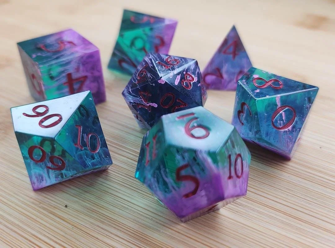 The photo shows JoaraCrafts the Atomic Swamp dnd dice set. A dice set with blue green see through body with purple and white tendrils coming from one of the sides. The numbers are colored in a pinkish red and the dice are set on a wooden background. 