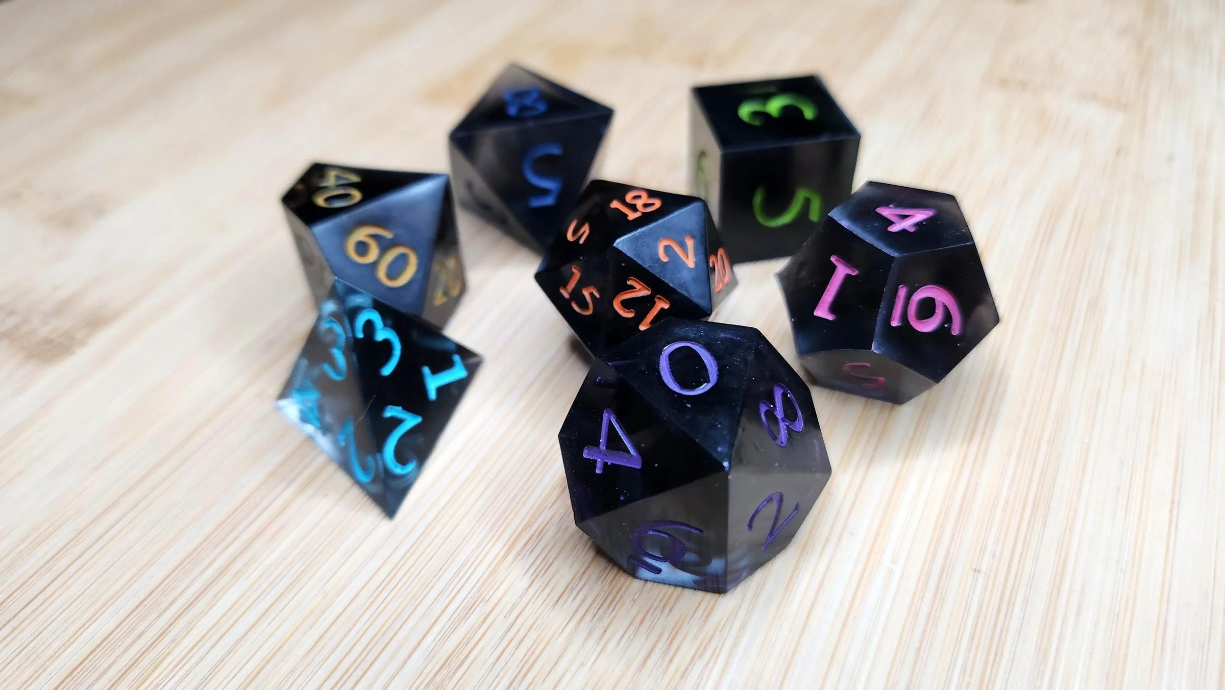 May include: A set of black polyhedral dice with rainbow colored numerals. The dice are in various shapes, including a d4, d6, d8, d10, d12, and d20. The dice are arranged on a light brown wooden surface.