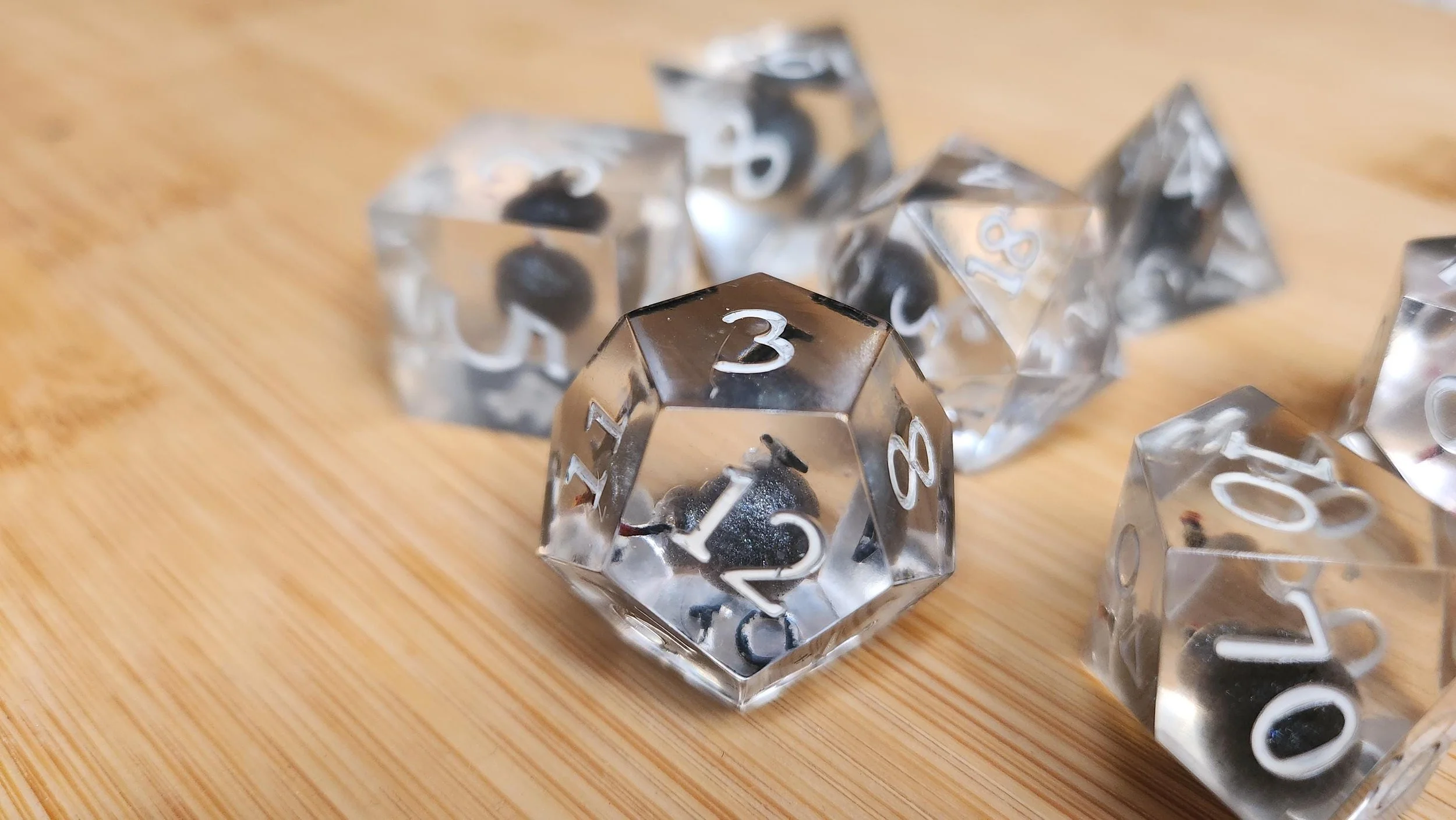 A detailed photo of the bombs away dice set with a focus on the 12 sided die. The white numbers color clearly visible over the clear dice and the floating ball bomb within. 
The dice are displayed on a wooden background.