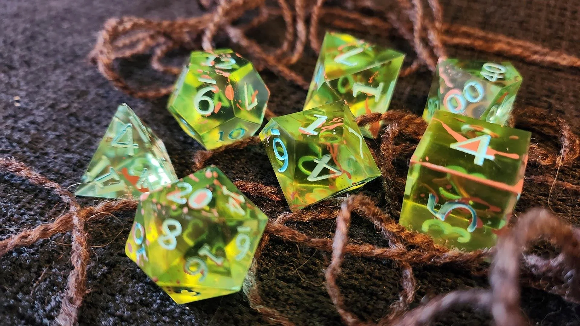 A detailed photo of the entangled dice with a focus on the 20 sided die. The blue numbers color clearly visible over the bright green and the brown spots covering the dice itself.
The background is dark with yarn acting as brambles