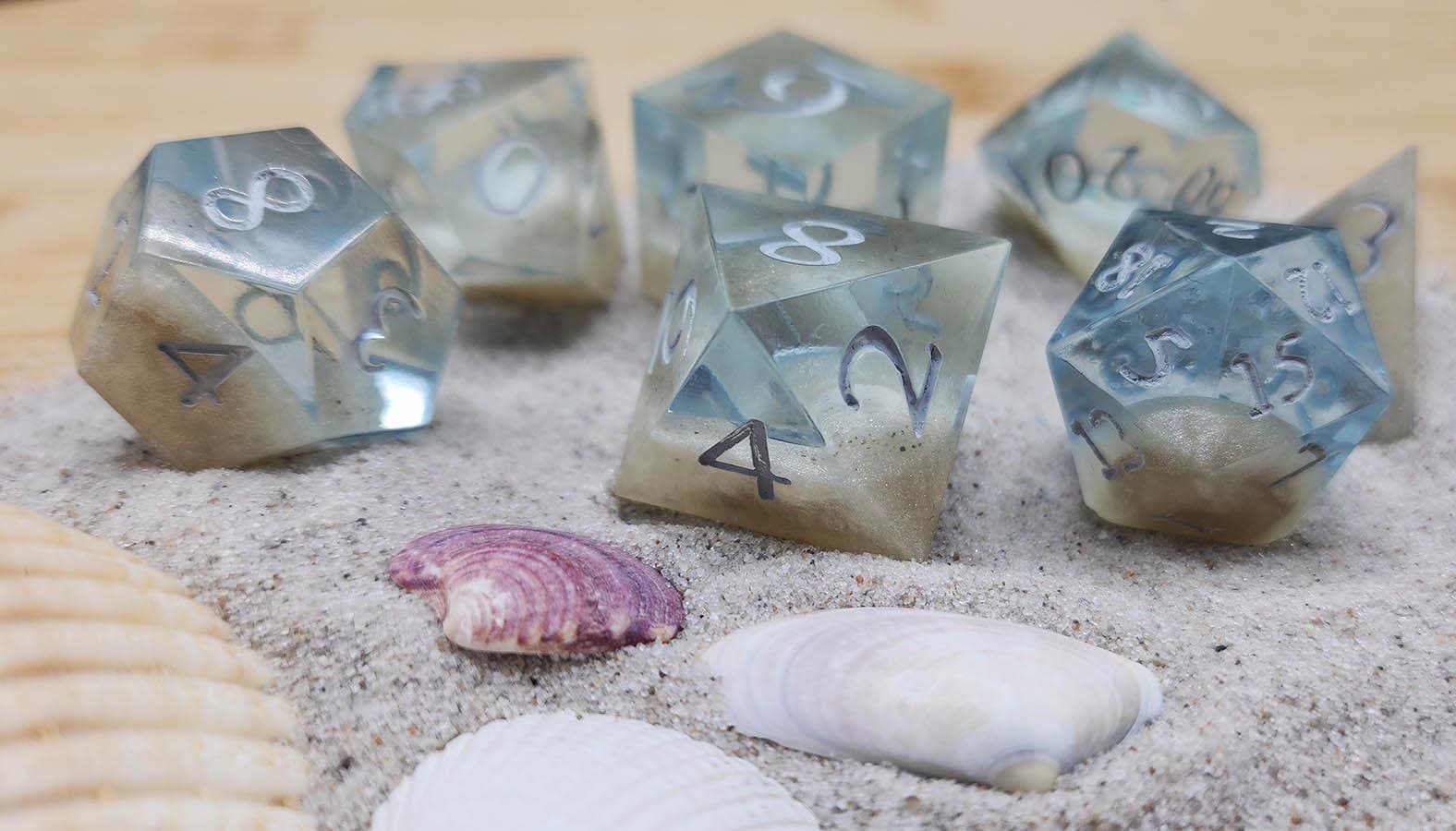 Joaracrafts Peaceful Ocean Dice Set for dungeons and dragons and other TTRPGs are a 7 piece dice set that mimic the appearance of real shells underwater. The dice are see-through blue and pale yellow with some sand to create a grainy effect, while th