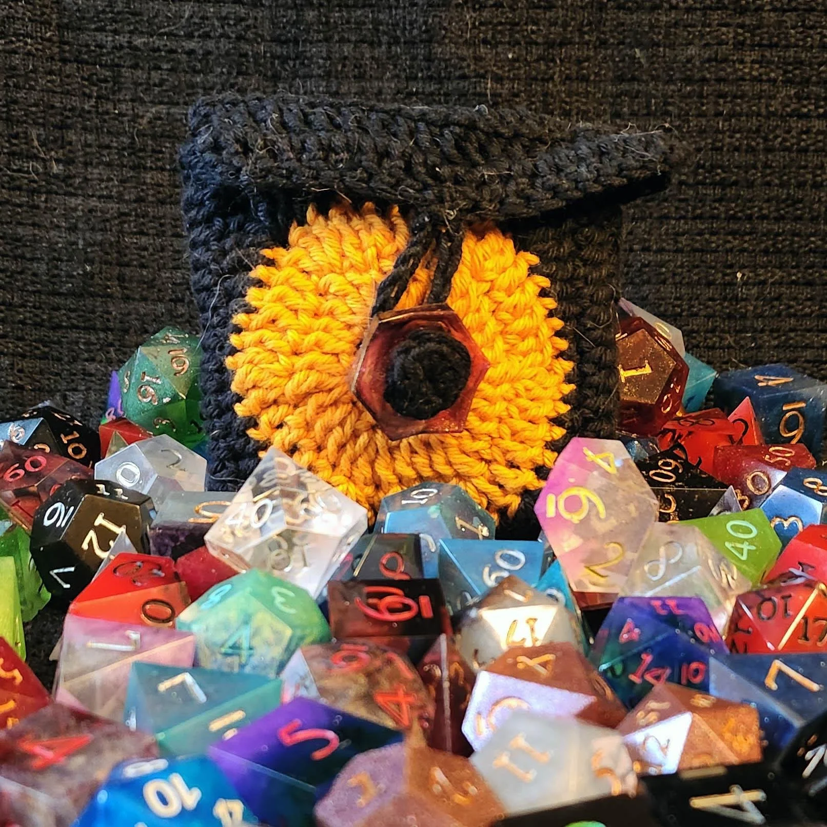 A photo of JoaraCrafts Anti-Waste Mystery Dice Pack. In this photo you can see a full handcrafted crochet black pouch with the orange hexagon representing the JoaraCrafts logo finished with a Anti-Waste resin button. The bag is then surrounded by sev