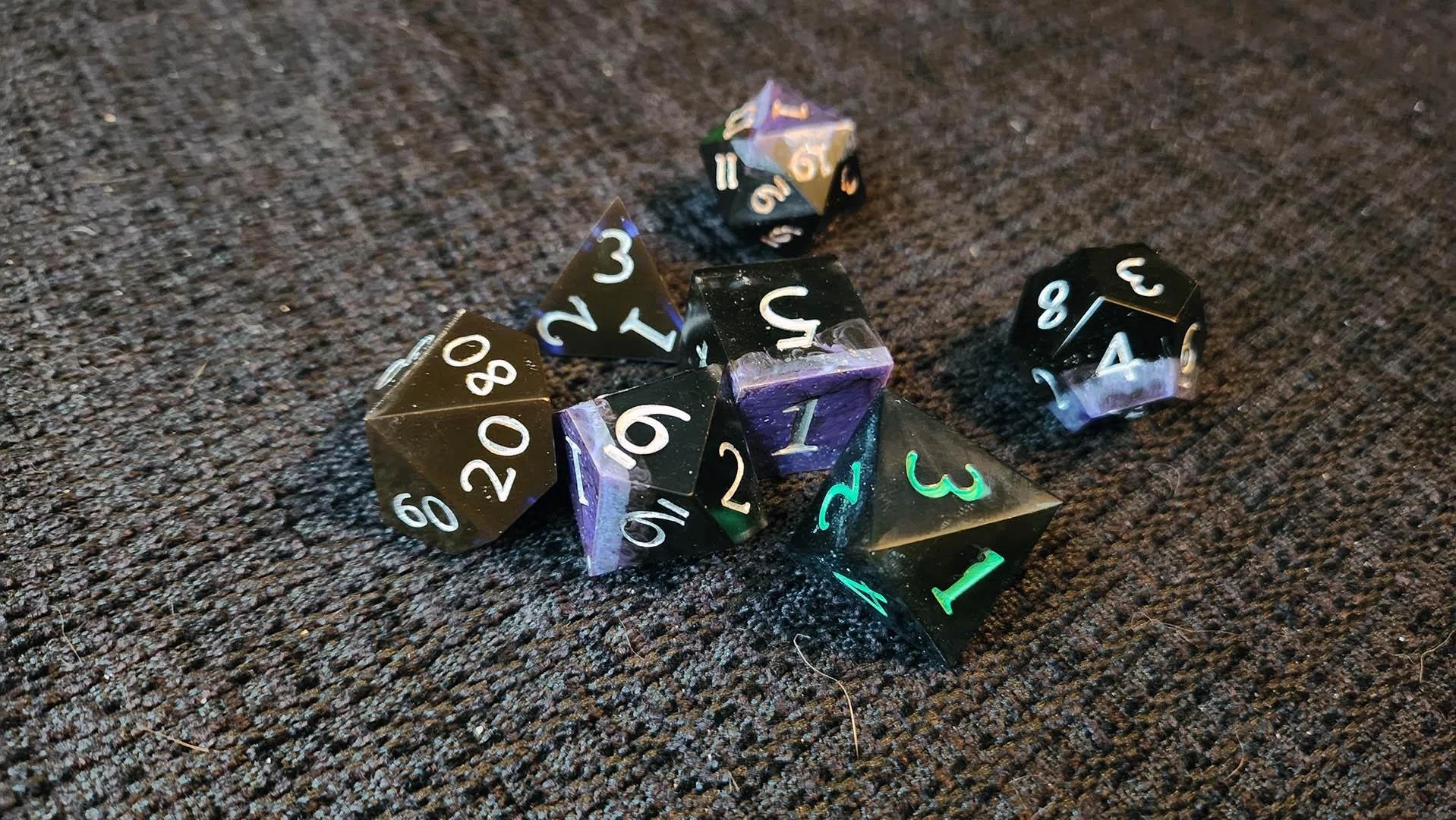 An example of how a JoaraCrafts mystery dice pack might look like. This specific set has a thematic black variant where all dice are mostly black, some  with some purple details and the numbers are either silver, gold or green.