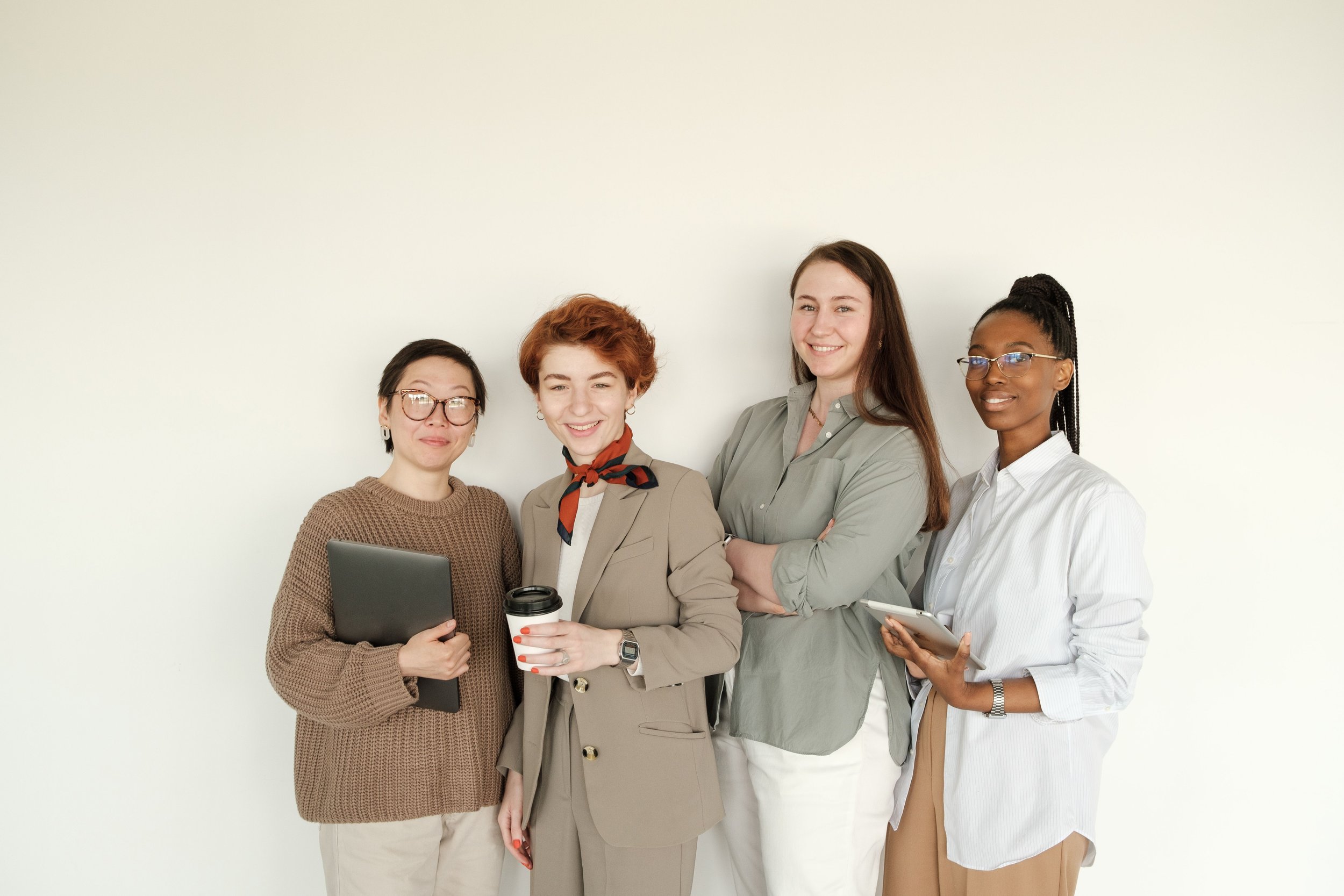 Group of five diverse women standing against a plain light-colored wall, smiling, holding tablets and coffee, dressed in business casual attire.