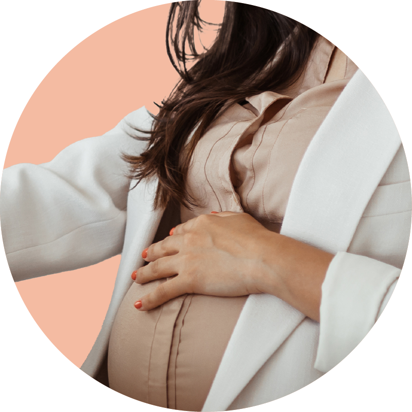 Close-up of a pregnant woman gently holding her belly with one hand, wearing a beige blouse and white blazer, against a pink background.