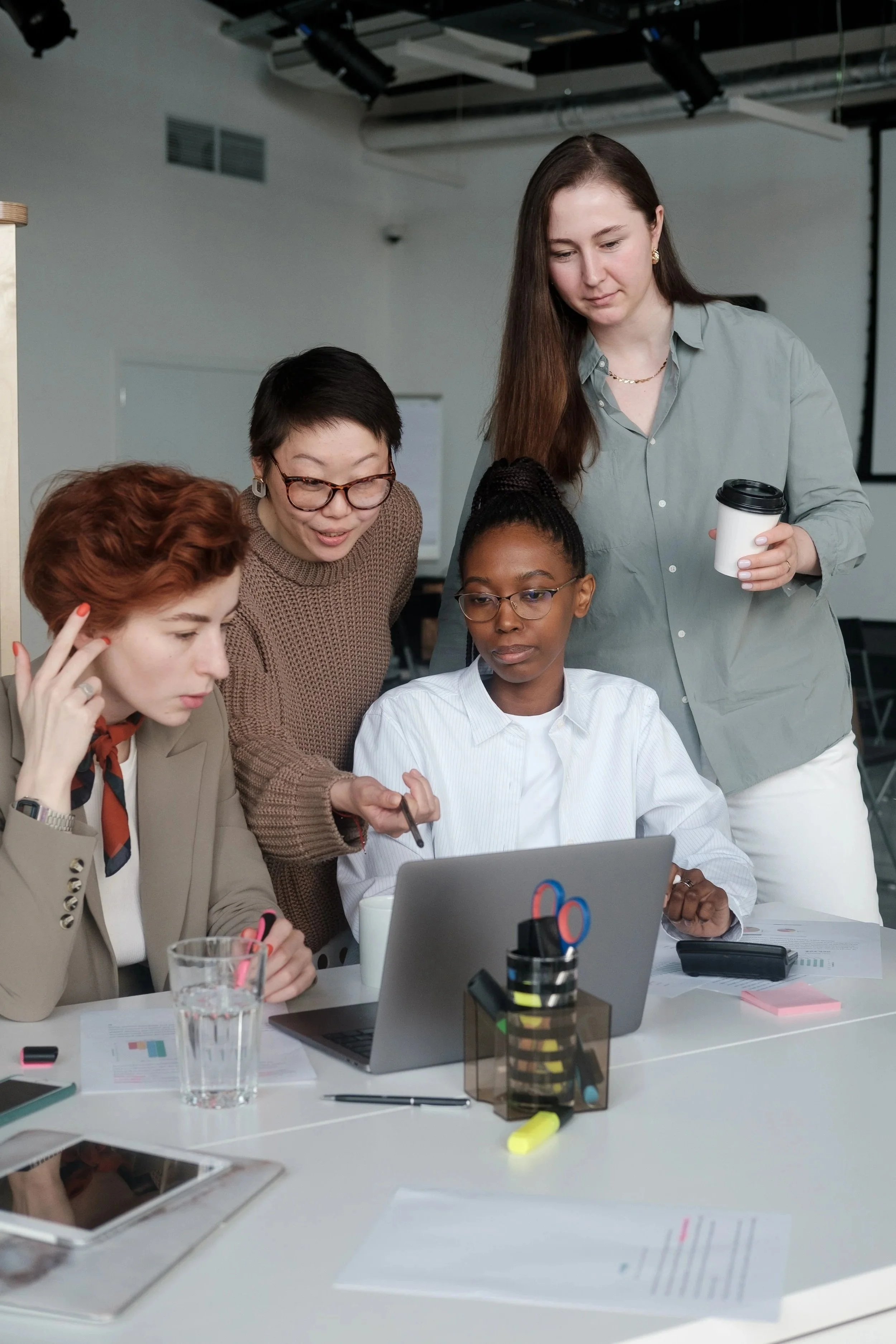 A diverse group of women working together in an office environment. They are gathered around a laptop, looking at the screen. Items on the table include documents, a glass of water, highlighters, and stationery. One woman is standing with a coffee cup, and others are seated, engaged in discussion.