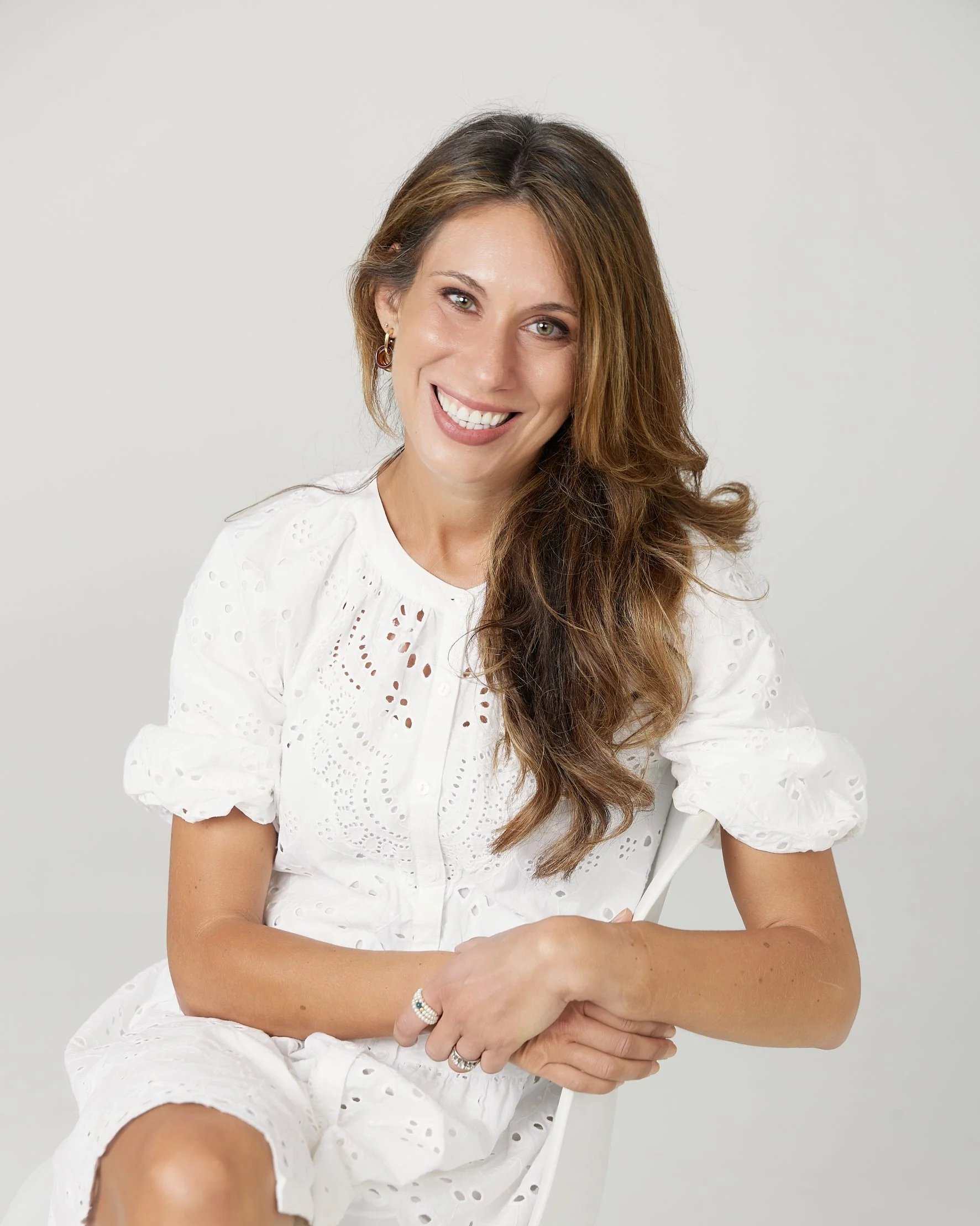 A woman with long brown hair smiling, wearing a white eyelet dress and rings, sitting against a plain gray background.