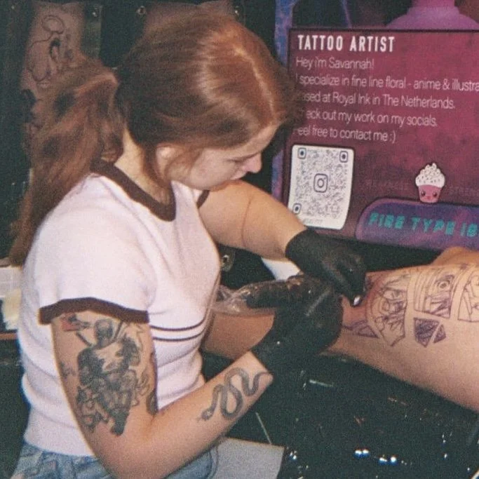 Tattoo-artist die een tattoo op een arm aanbrengt, met tattoos zichtbaar op haar eigen arm, achter haar een poster met informatie over haar als tattoo-artist.