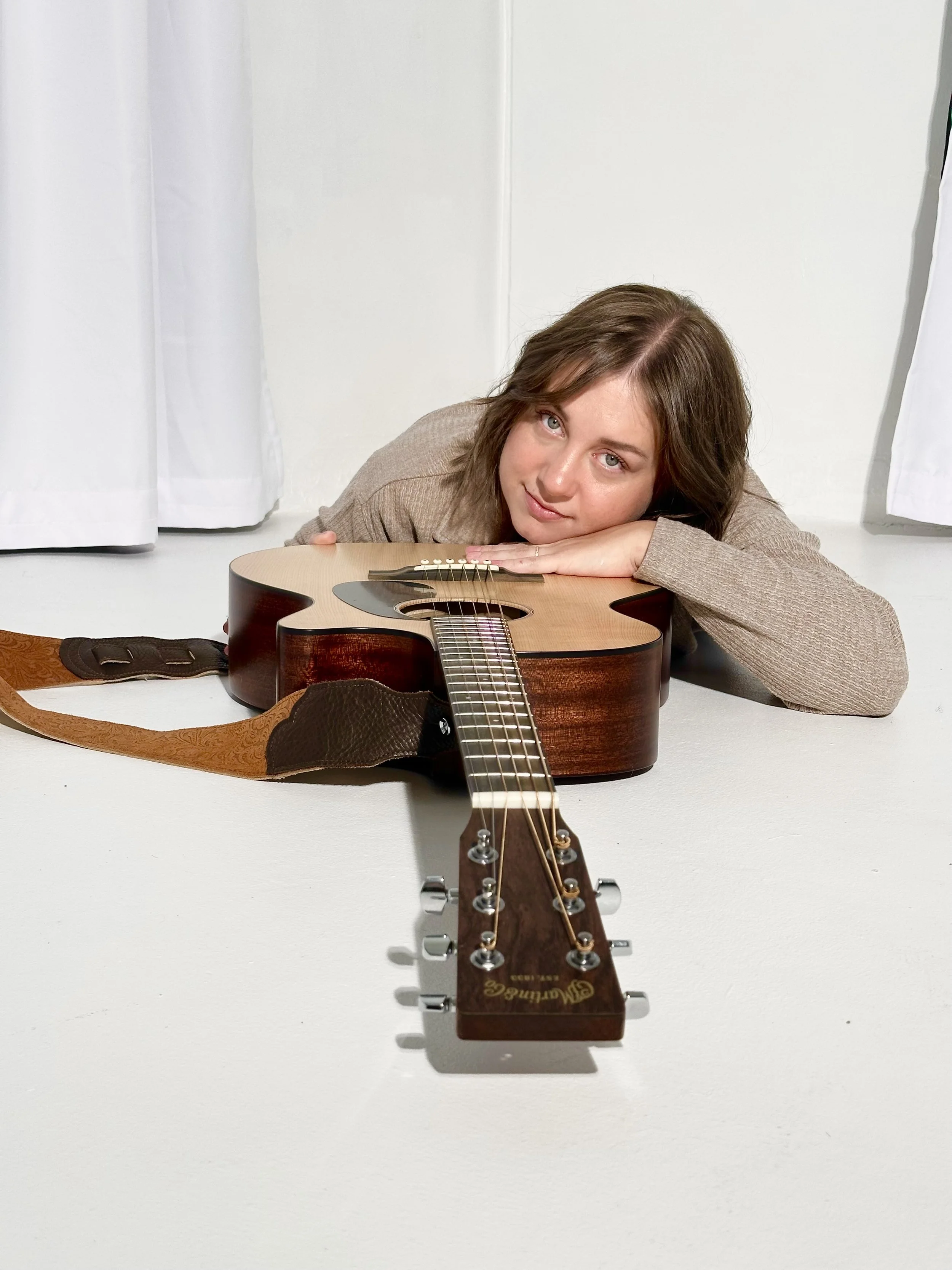 Emmy Jessen resting her head on her arms behind an acoustic guitar in a bright studio setting.