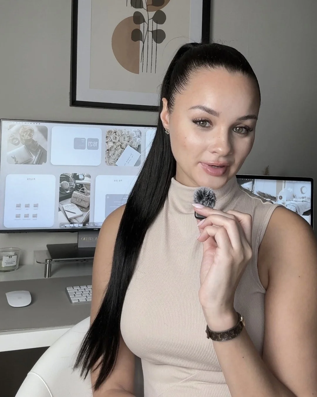 Een vrouw met zwart haar in een paardenstaart houdt een microfoon vast en kijkt in de camera, op de achtergrond bevinden zich computerschermen met diverse foto's en een abstract kunstwerk aan de muur.