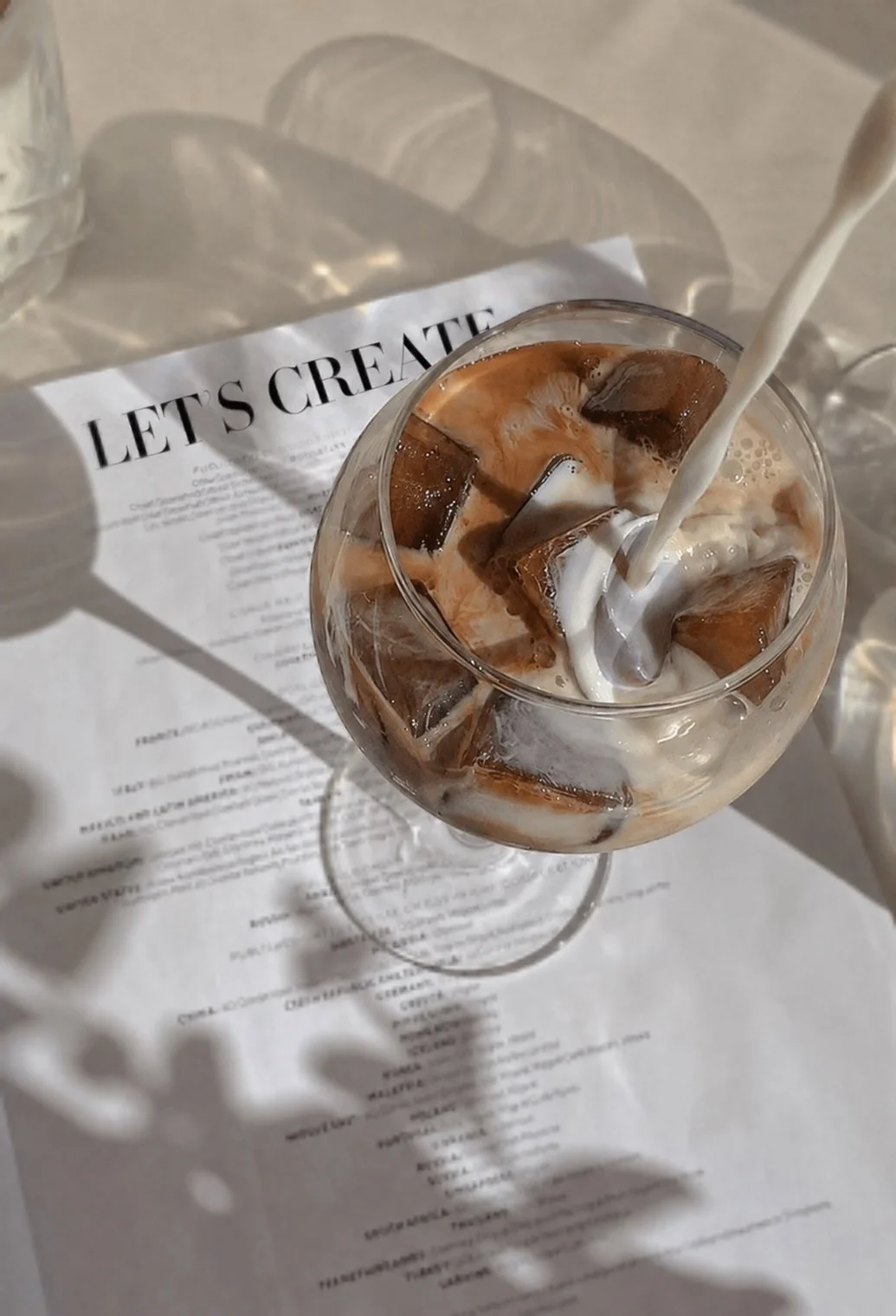 Een glas met ijsklonten, room en chocola, met een vloeistof die wordt ingeschonken, op een tafel met een papieren menu met de titel 'LET'S CREATE' en een schaduw van een bloem.