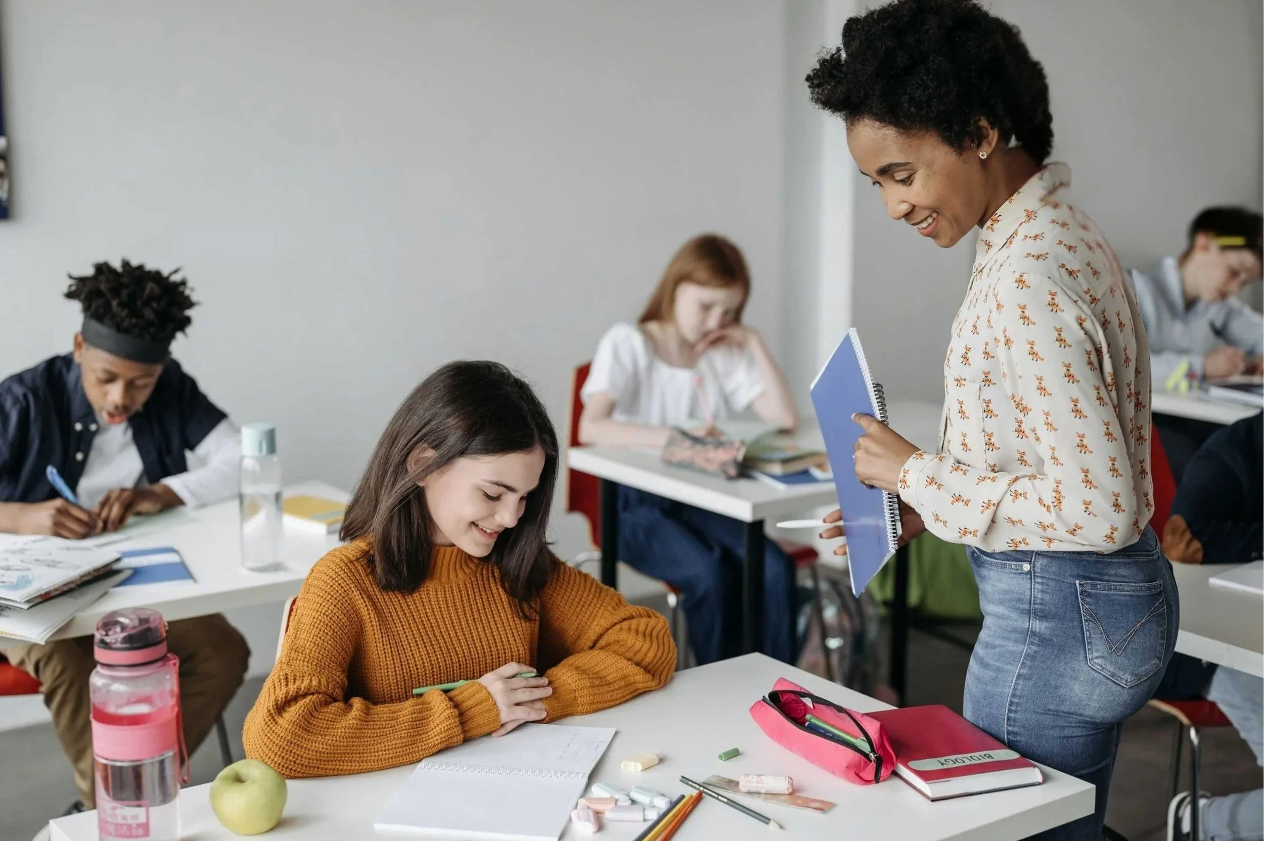 A classroom scene with a smiling female teacher engaging with students. She holds a blue notebook, leaning over a student who is writing in a notebook. The students are seated at desks with school supplies, notebooks, and water bottles. The classroom background is plain and bright.