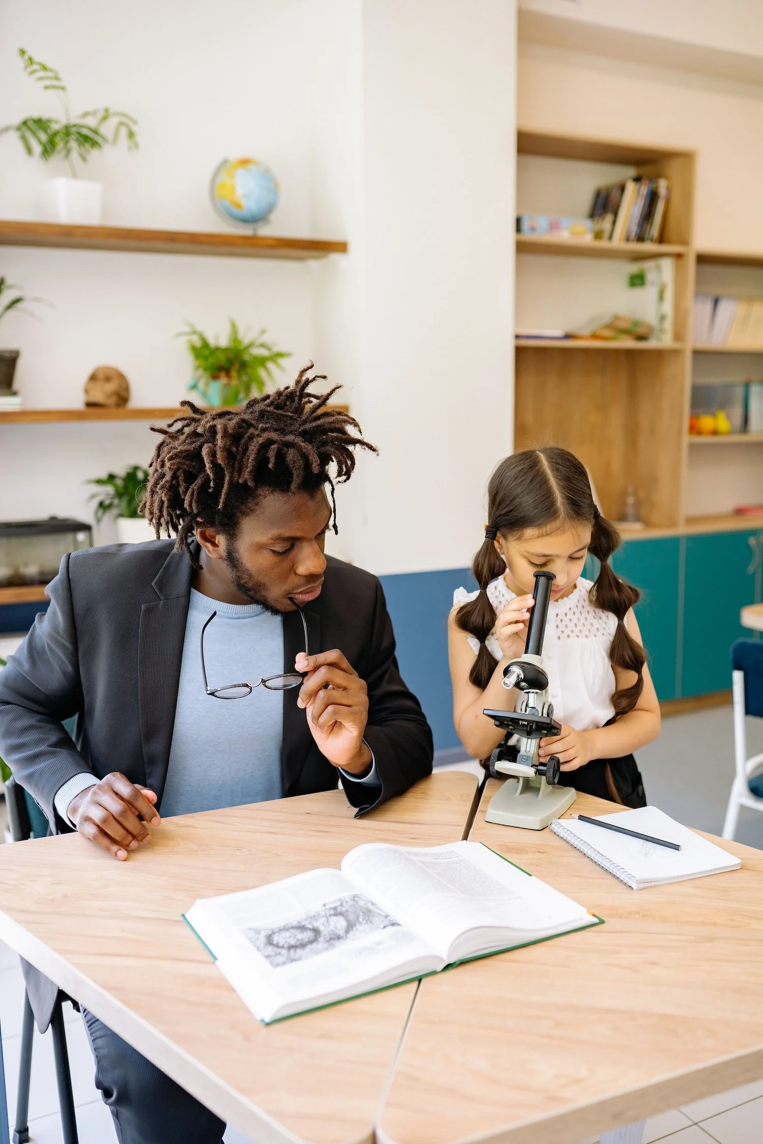 A young girl with pigtails using a microscope while an adult man looks on, at a desk with open books and notebooks, in a classroom or science lab setting.