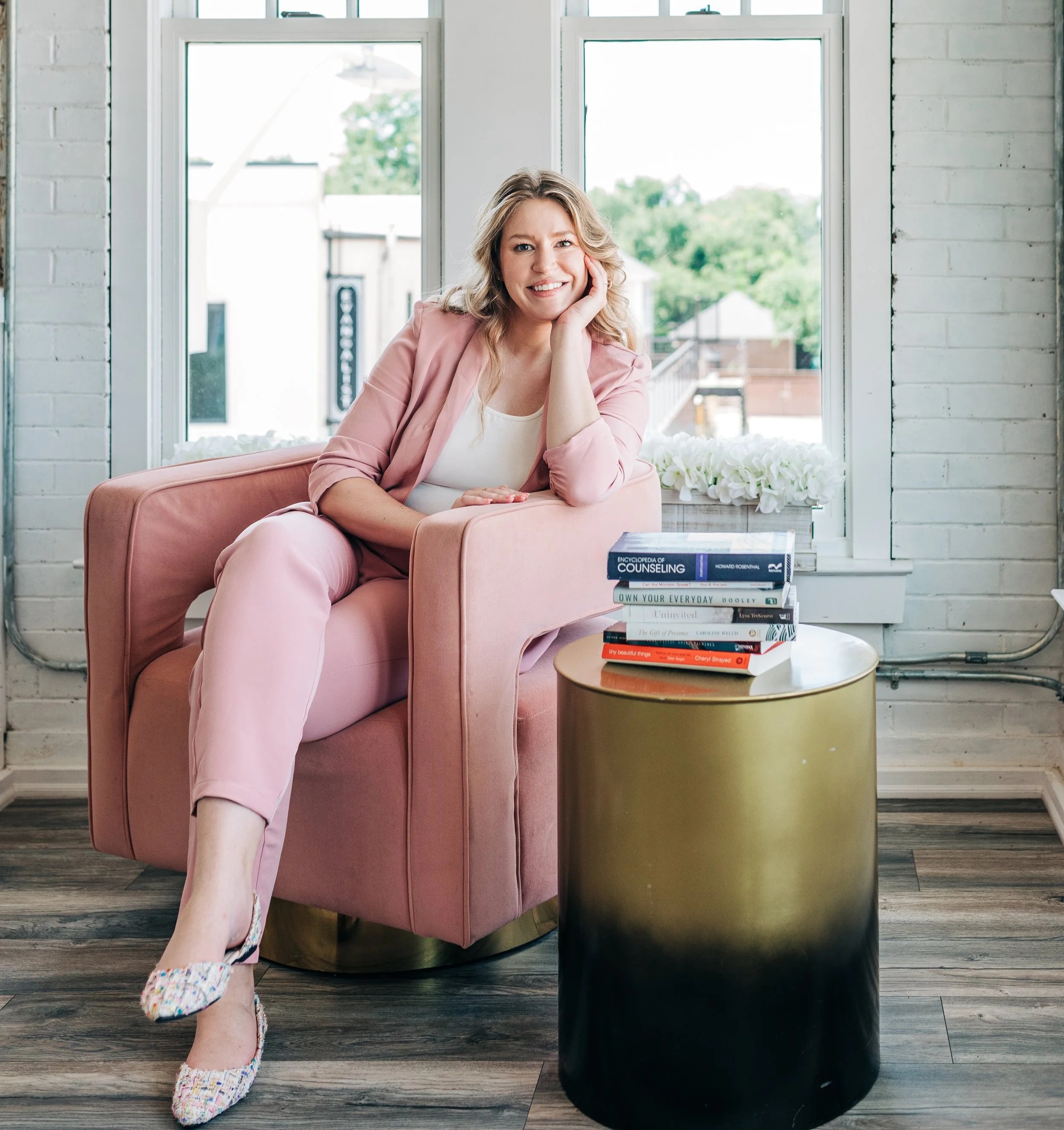 A woman with blonde hair smiling, sitting on a pink armchair in a room with white brick walls and large windows, wearing light pink pants, a white top, a pink blazer, and colorful woven slippers. There is a stack of books on a round side table next to her.