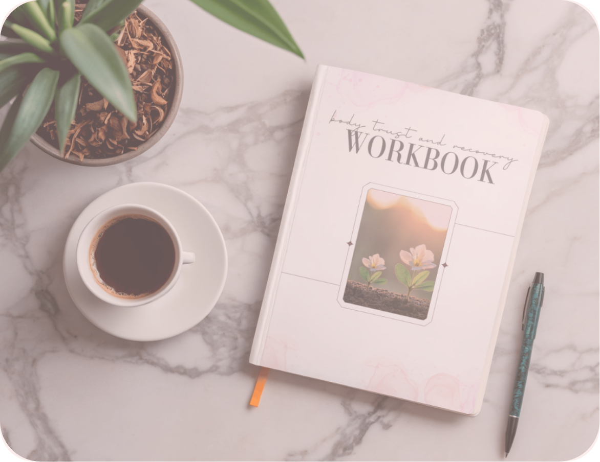 A white marble desk with a potted green plant, a cup of black coffee on a saucer, a white recovery workbook titled 'Body Trust and Recovery Workbook' with an image of two flowers, and a blue-patterned pen.
