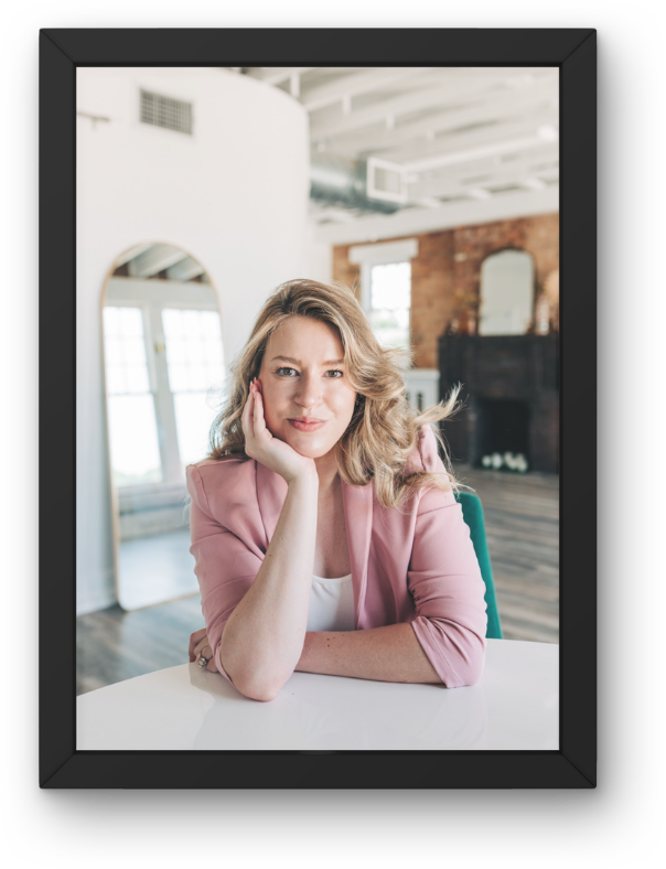 A woman with wavy blonde hair, wearing a pink blazer and white top, sitting at a white table indoors, resting her face on her hand, smiling at the camera. The background features large windows, a mirror, brick wall, and a fireplace.