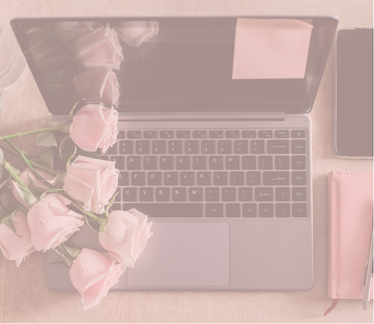 A laptop with a black keyboard and silver body on a light wooden surface, surrounded by pink roses, a pink notebook with a pen, and a smartphone.