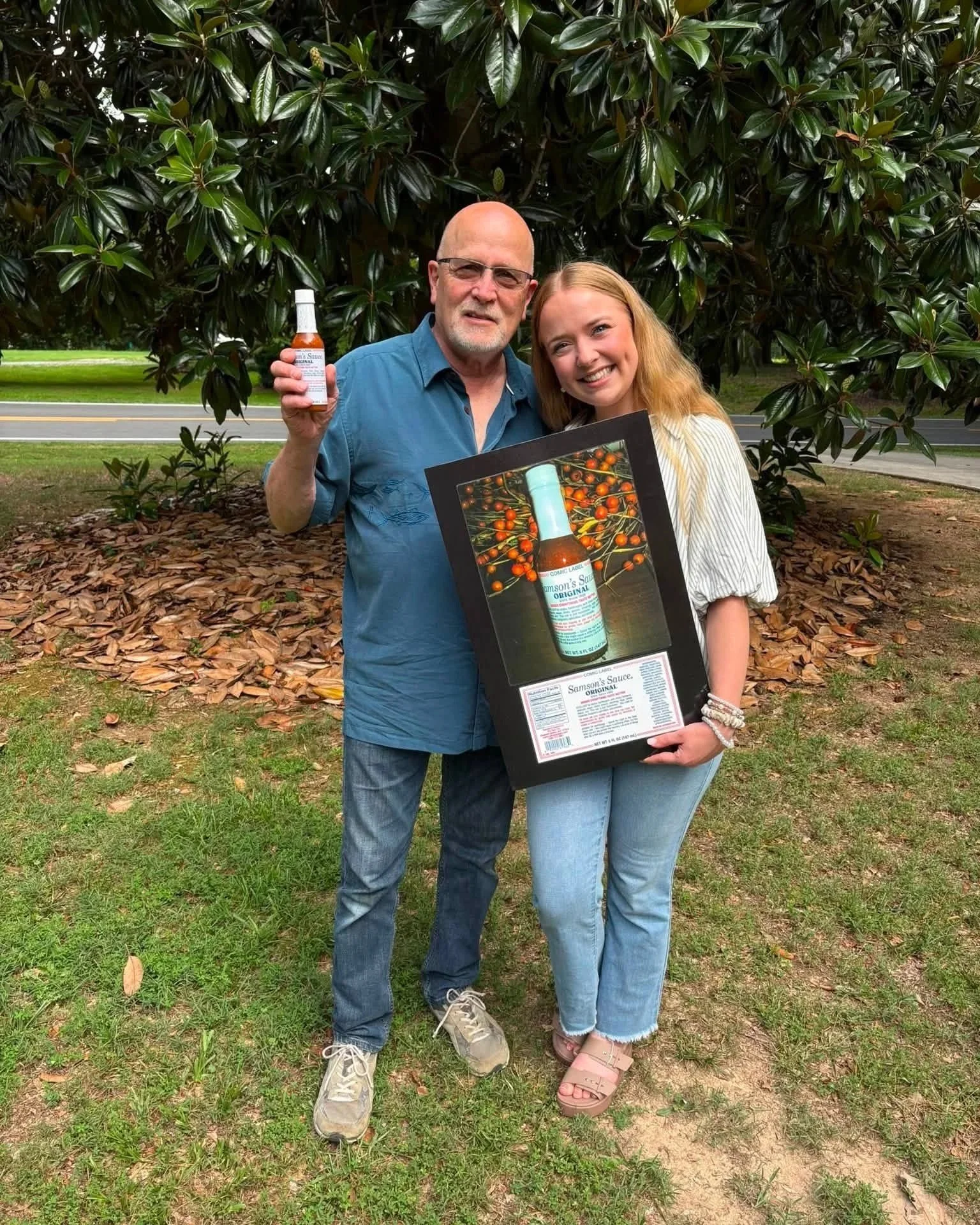 A man and a young woman standing outdoors in front of a large leafy tree, smiling at the camera. The man is holding a small bottle of hot sauce, and the young woman is holding a framed picture of a larger bottle of hot sauce with a background of small orange peppers.