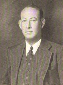 Black and white portrait of a man dressed in a suit with a tie, posed against a plain background.