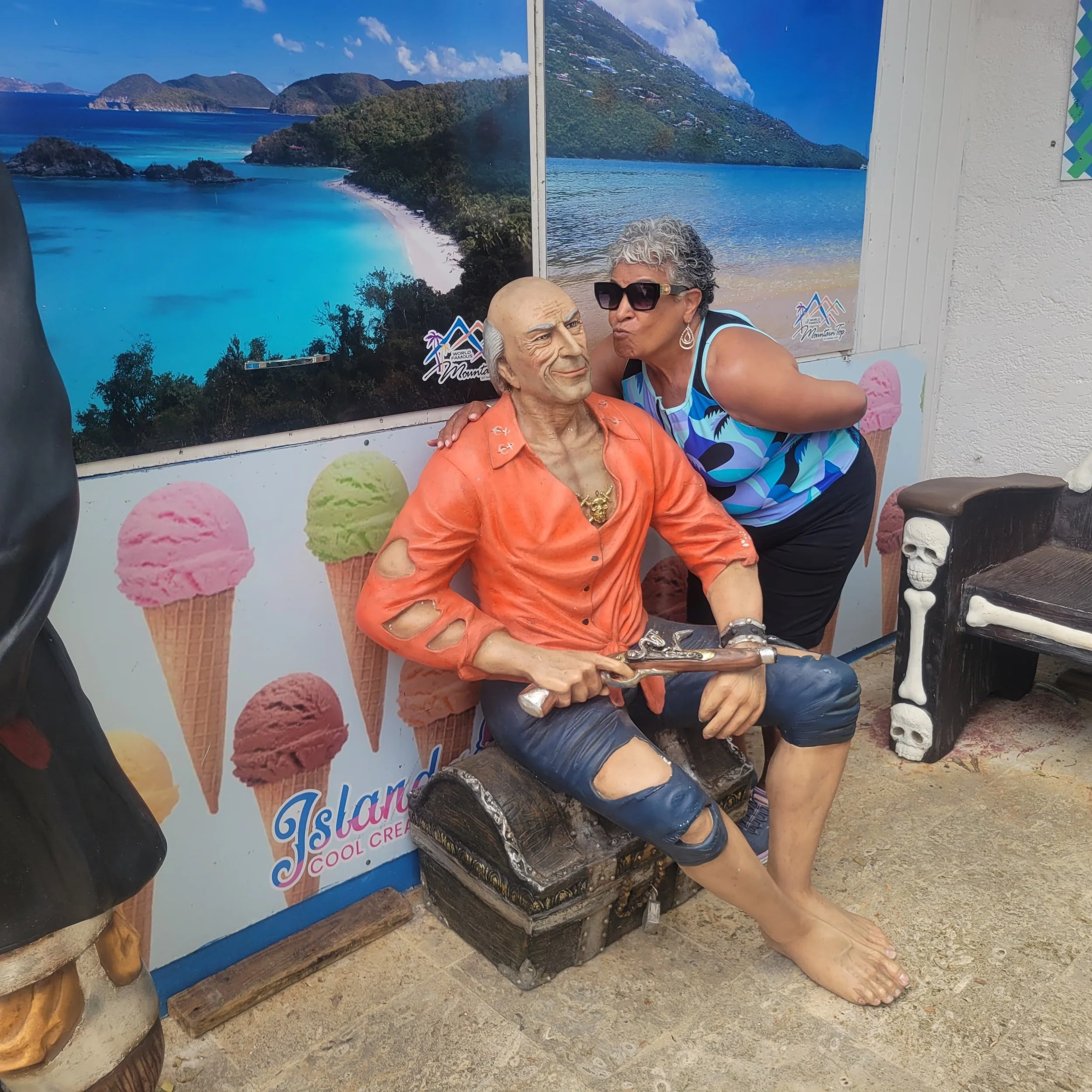 A woman with short curly gray hair wearing sunglasses and a colorful dress posing next to a wax figure of an older man, with a tropical island beach scene in the background.