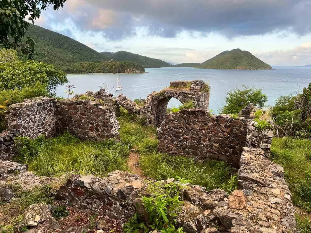 Ruins of old stone structures on a grassy hill overlooking a body of water with sailboats, surrounded by green hills and distant islands under a cloudy sky.