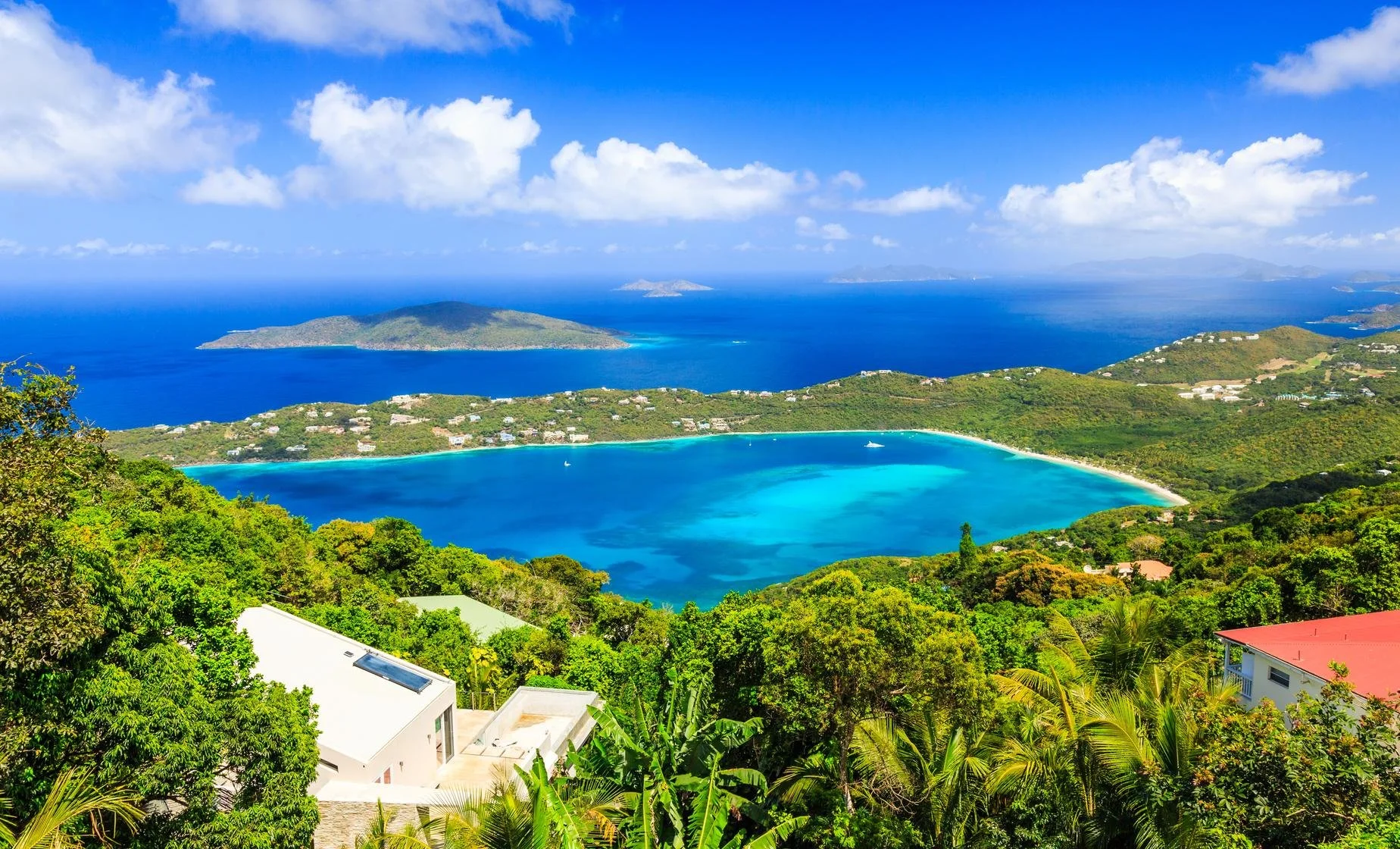 Aerial view of a tropical coastal landscape with a blue bay, lush green hills, and distant islands under a partly cloudy sky.
