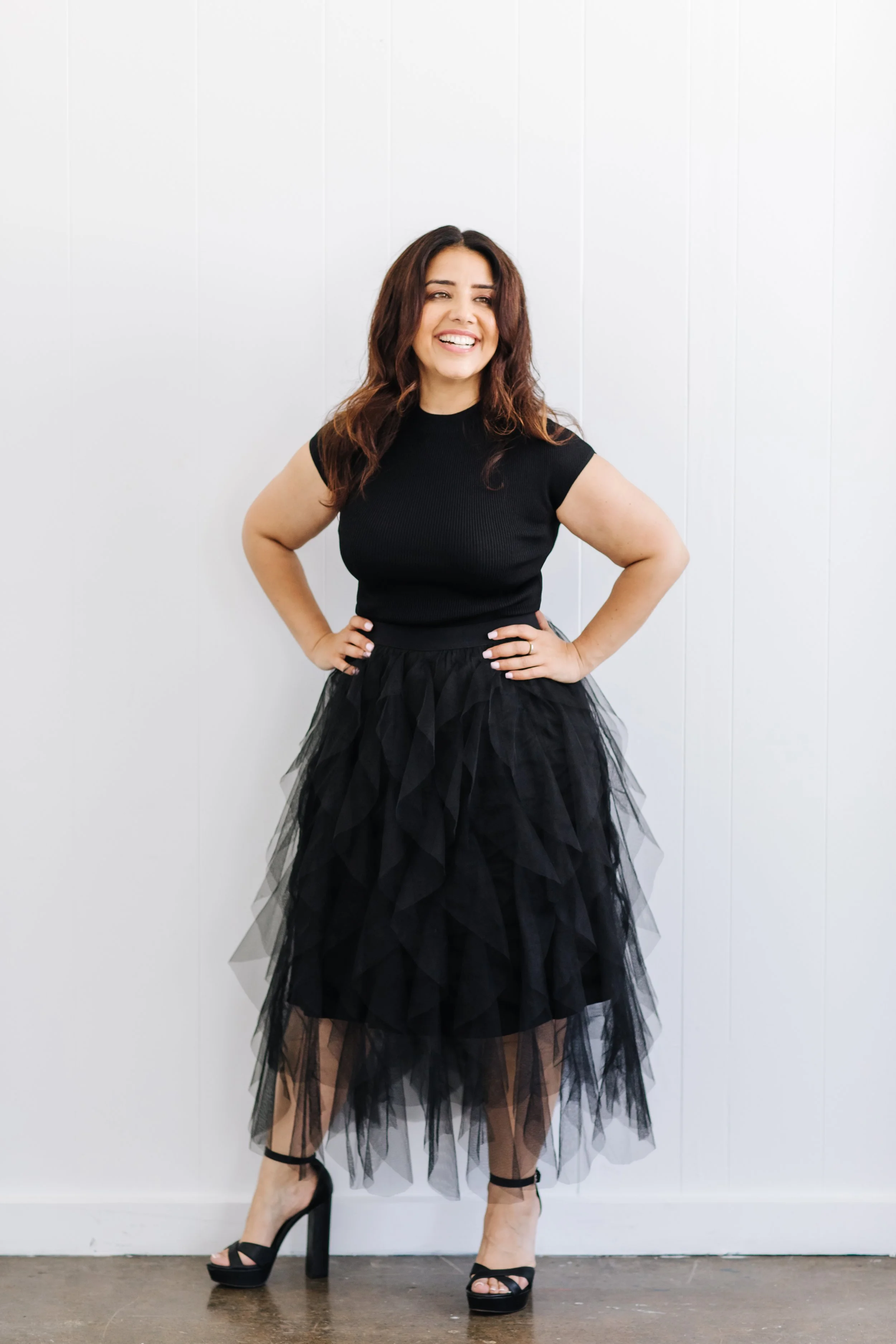 Woman in black sleeveless top, black tulle skirt, black high heels, standing against a white wall, smiling with hands on hips.