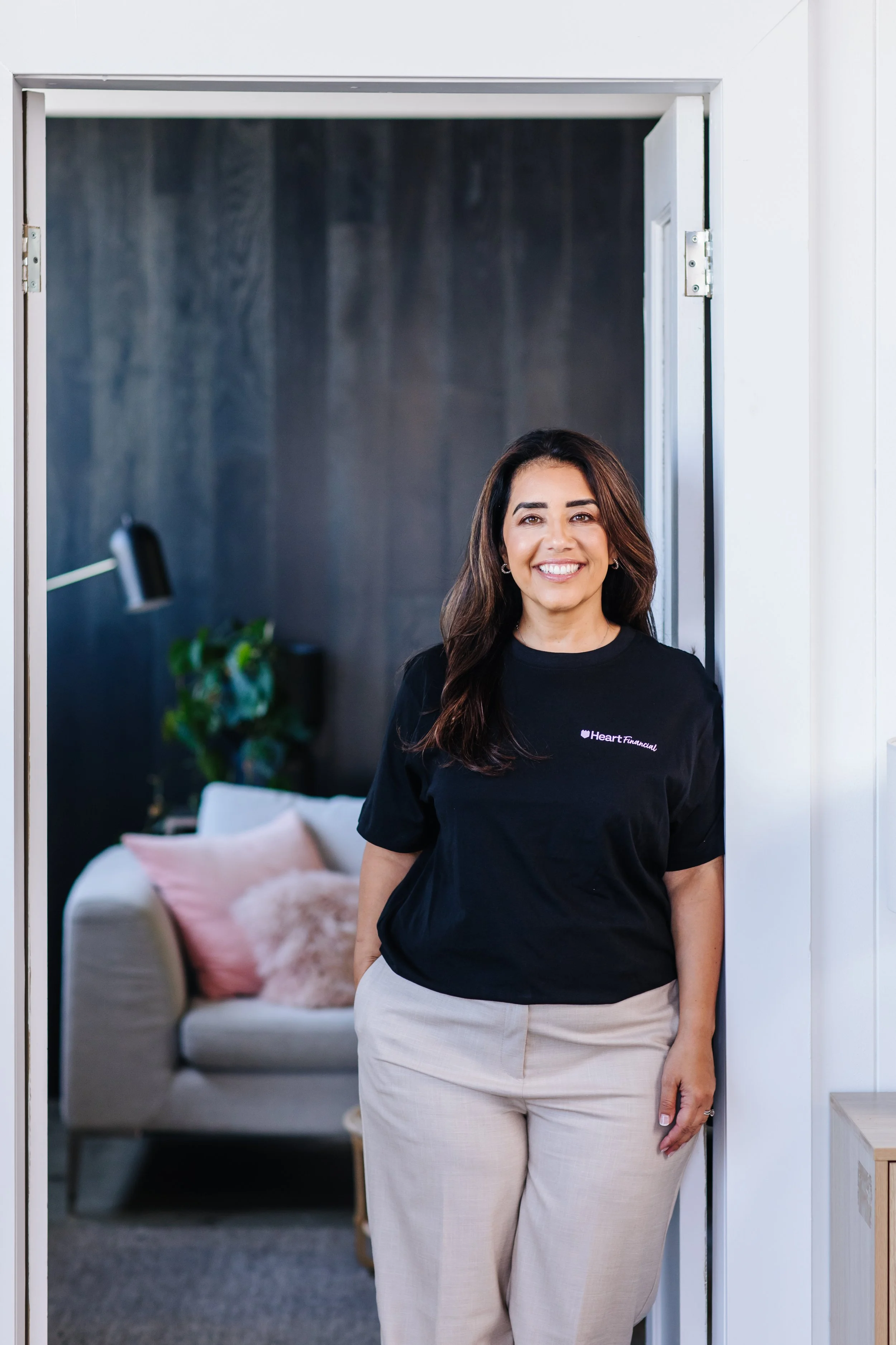 A woman with long brown hair smiling and standing in a doorway, wearing a black t-shirt with white pants.