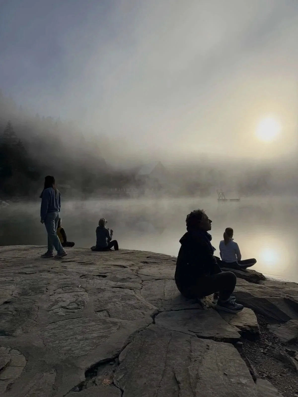 People sitting and standing on a rocky shore by a foggy river or lake at sunrise, with mist and a boat in the distance.