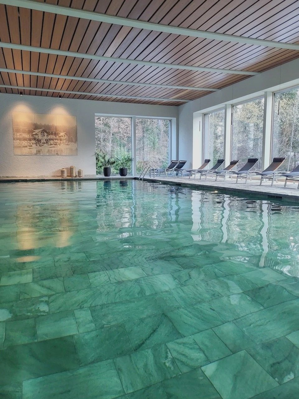 Indoor swimming pool with greenish tiles, surrounded by lounge chairs and large windows showing an outdoor view of trees and nature.