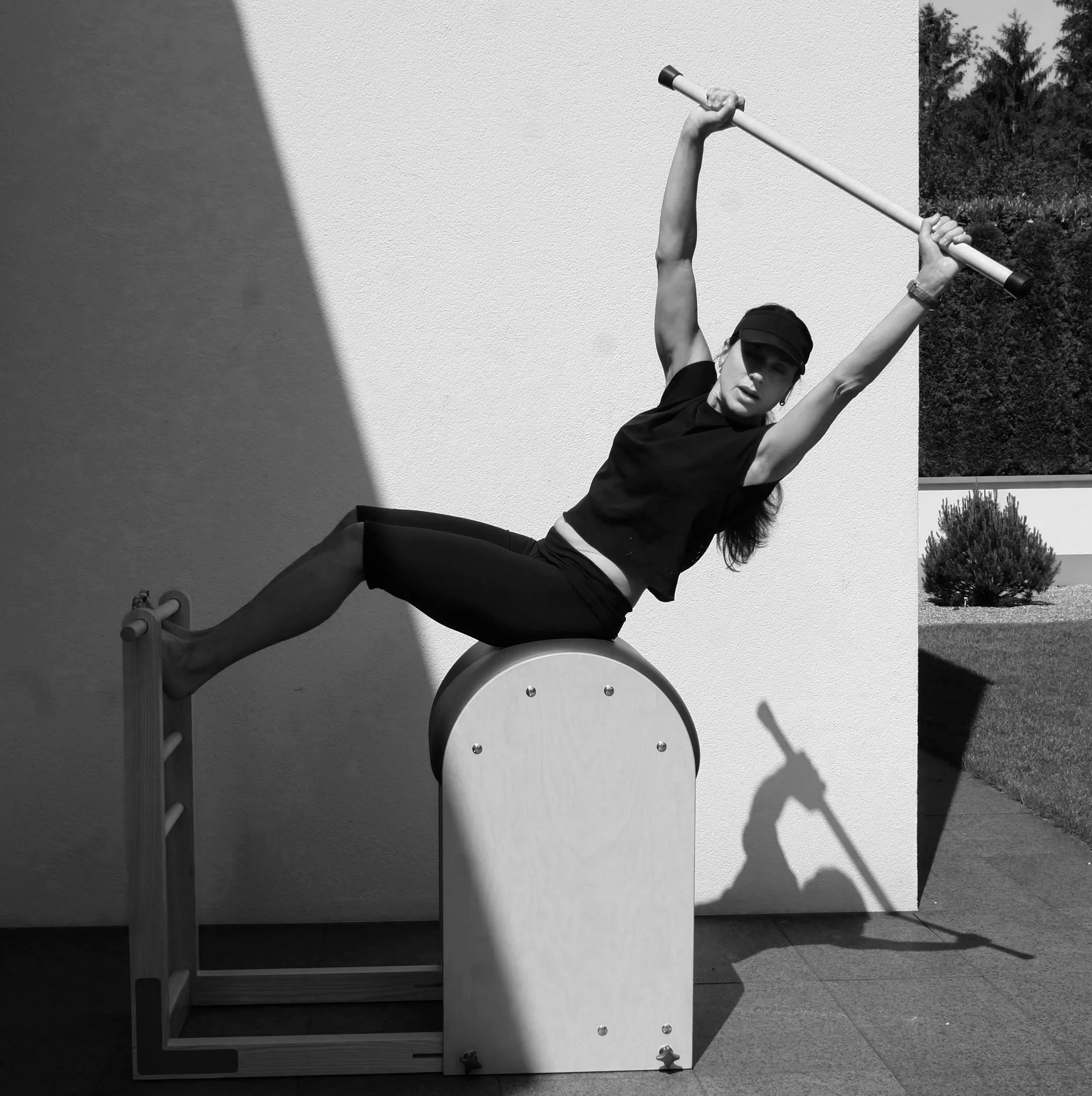 A woman performing a yoga pose outdoors against a white wall, wearing dark clothing and a cap, with palm trees and sky in the background.