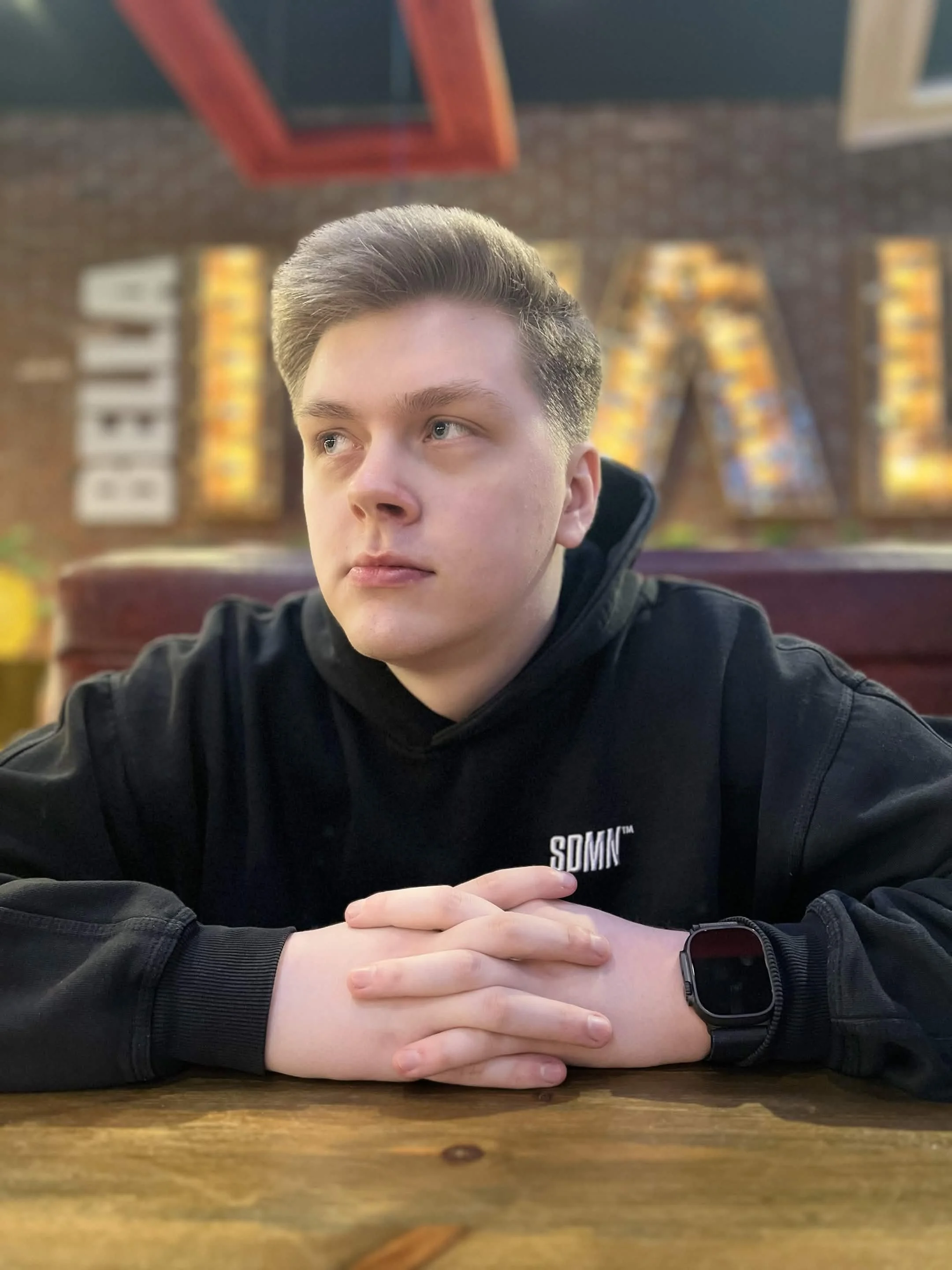 A young man with styled hair sitting at a wooden table inside a restaurant. He is wearing a black hoodie with the text SDMN and a smartwatch on his left wrist. His hands are clasped together on the table, and he is looking to the side. The restaurant has a brick wall and colorful framed murals in the background.