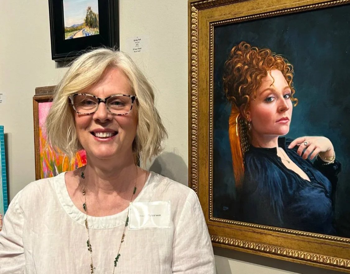 An artist woman with blonde hair and a white top standing next to a framed portrait painting of a woman with curly red hair wearing a black top in a traditional style.
