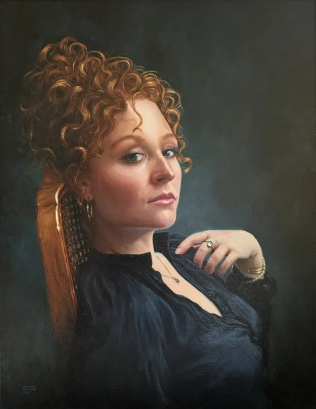 A woman with curly red hair sitting on a chair, wearing a black top, gold hoop earrings, a ring, and a necklace, looking over her shoulder at the camera with a confident expression.